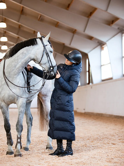 A woman in a Uhip arctic puffer coat extended  eclipse  and black boots stands in an indoor horse stable next to a dapple grey horse, holding its reins.