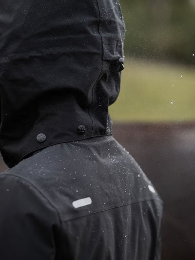 Close-up of a person wearing a  black Uhip Rainy Jacket outdoors, with raindrops visible on the fabric and blurred greenery in the background.