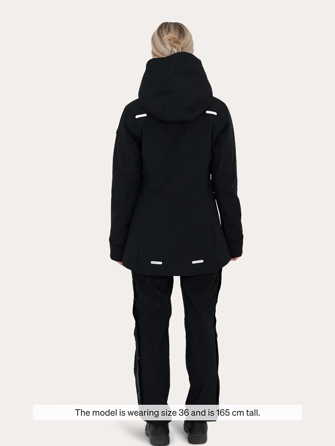 A model viewed from the back wearing a  black Uhip Rainy Jacket and matching black ski pants against a light grey studio background. A text overlay at the bottom reads, 'The model is wearing size 36 and is 165 cm tall.
