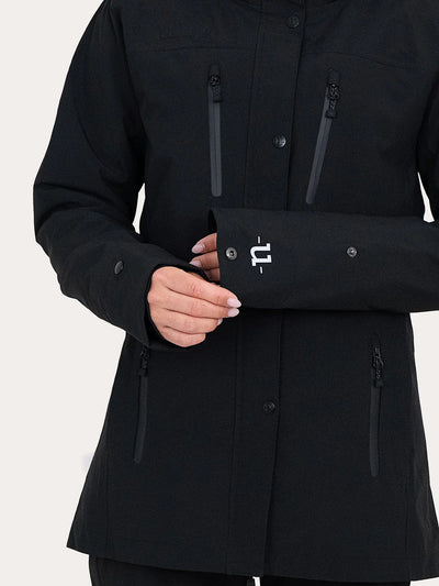 A close-up of a person wearing a black Uhip Rainy Jacket showing the front pockets, buttons, and a white stylized logo on the cuff, against a light neutral background.