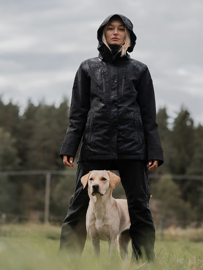 A woman wearing a Uhip Rainy  Jacket black waterproof hooded jacket and black pants stands in a grassy field with a light-colored Labrador Retriever dog under an overcast sky, with trees in the background.