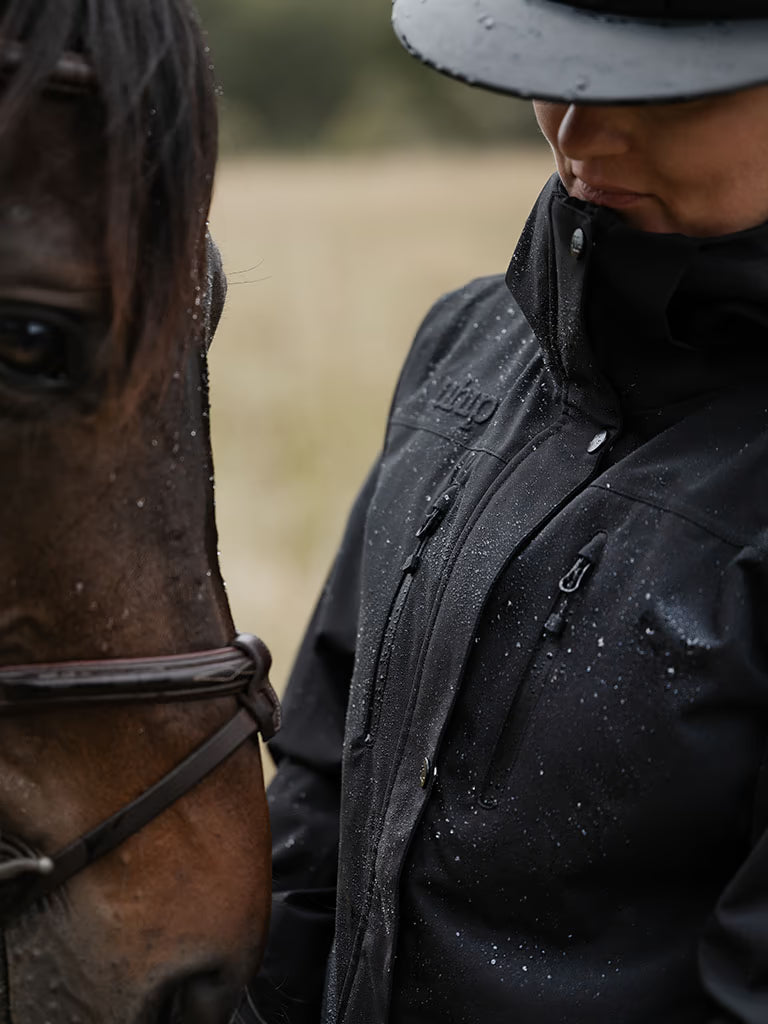 A close-up outdoor shot of a person in a Uhip Rainy Jacket black waterproof riding jacket and a grey riding hat standing next to a horse with water droplets on their clothing, against a blurred field background.