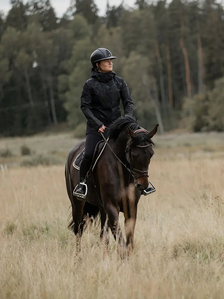 A person in a black Uhip Rainy Jacket  sits on a dark horse in a grassy field with trees in the background.