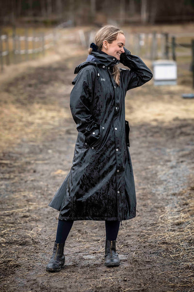 A woman wearing a Uhip Winnie  Raincoat jet black with black leggings, and black boots stands on  a muddy outdoor path.