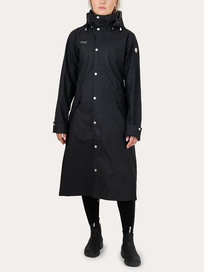 A model wearing a Uhip Winnie  Raincoat jet black   with silver snap buttons, black pants, and black boots, standing against a light gray studio background.