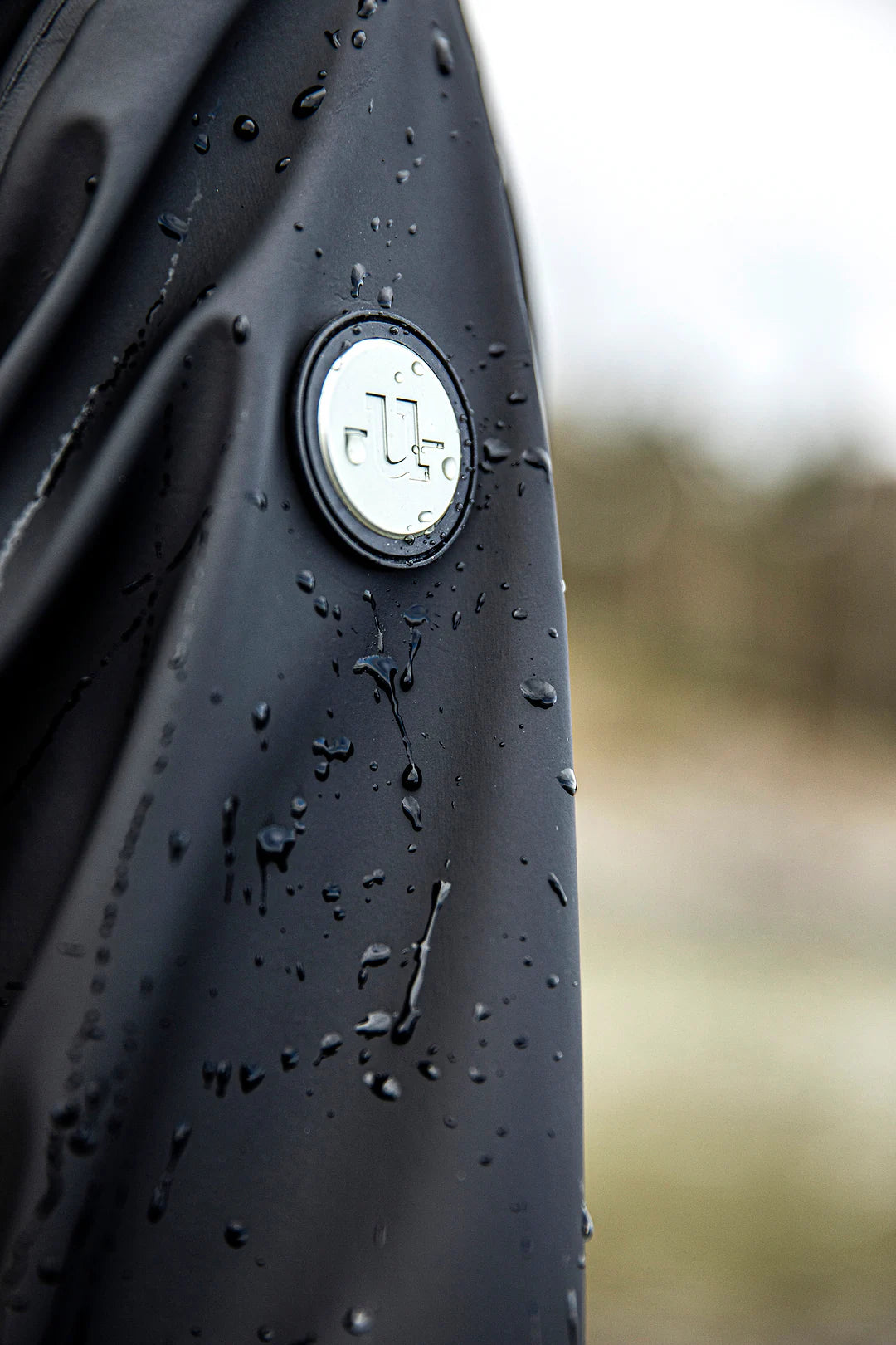 Close-up of a  Uhip Winnie  Raincoat jet black covered in water droplets, featuring a circular silver and black Uhip logo badge on the chest, with a blurred outdoor background.