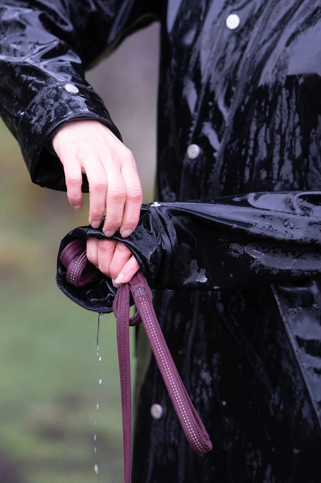 A close-up of a person wearing a Uhip Winnie Raincoat jet black in the rain, holding the cuff of the coat to show a dark burgundy dog lead and strap detail.